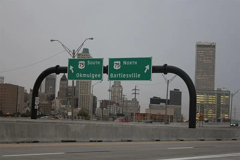 An overhead highway sign structure of bent steel pipe spanning US-75 in Tulsa, with the downtown skyline behind it